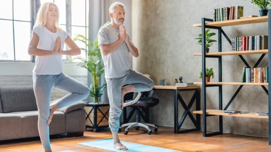 A senior couple is seen standing in tree pose on their yoga mats in the living room-4.png