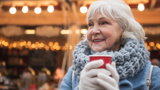 A senior woman wearing gloves and a scarf drinks a hot beverage outside-1.png
