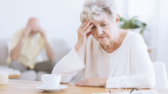 An older woman sits at a table with coffee looking distraught, with a man in the background with his head in his hands-6.png