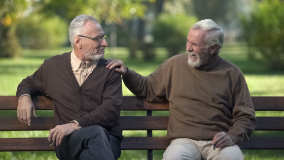 Two men in sweaters sit on a bench at the park talking and smiling-7.png