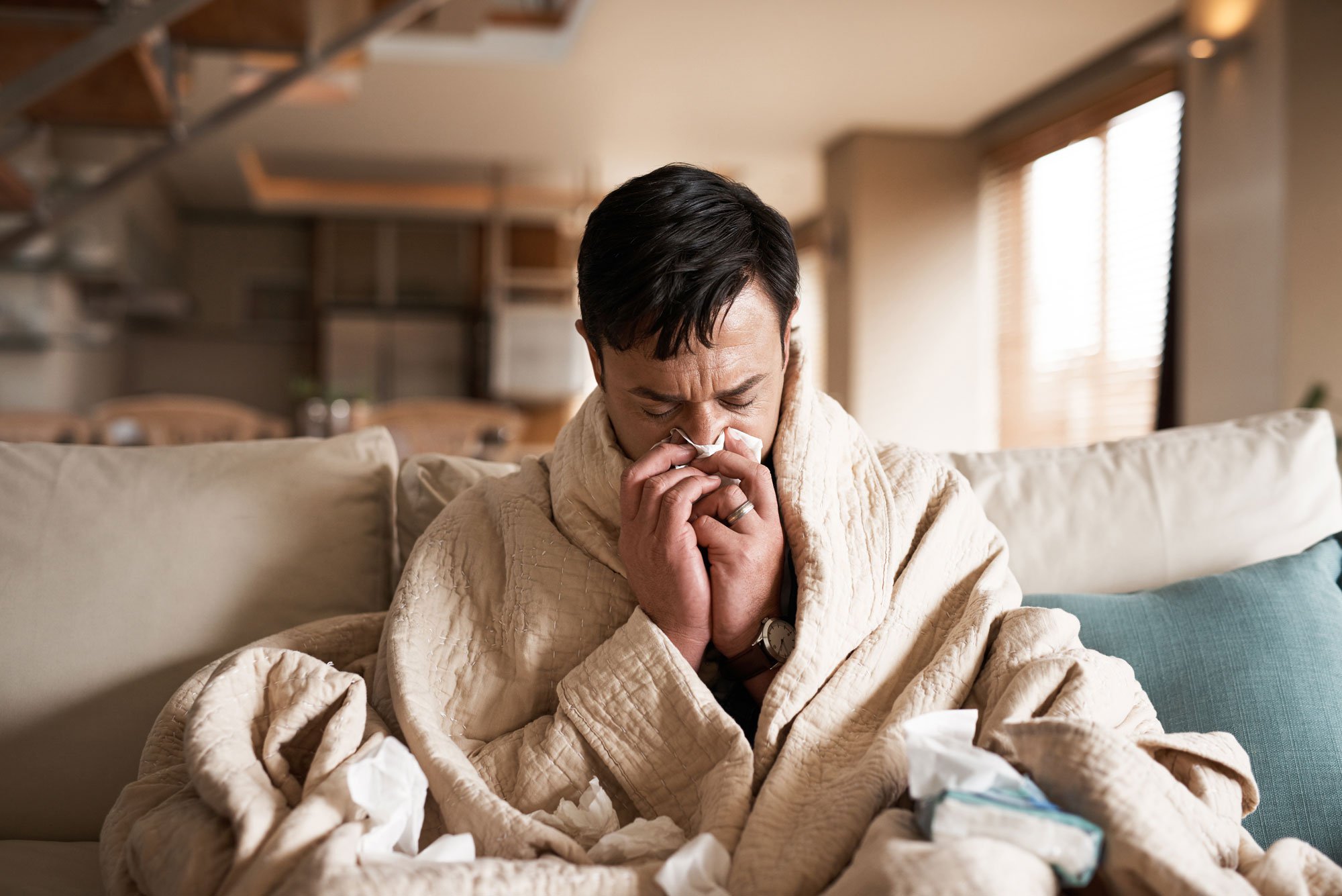 Man sitting on couch with a blanket wrapped around him, blowing his nose