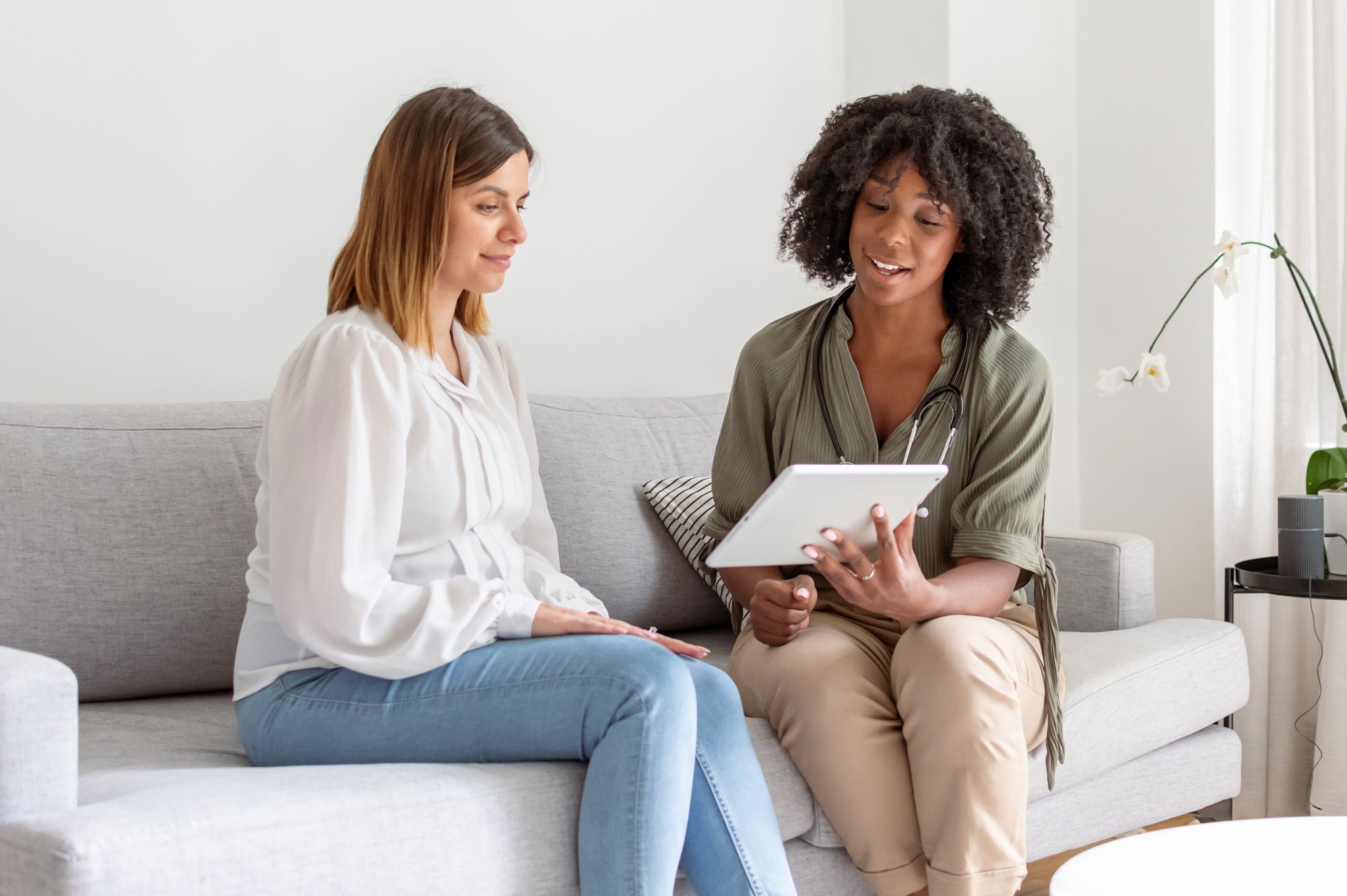 Female doctor sitting next to a female patient on the couch showing her something on a tablet