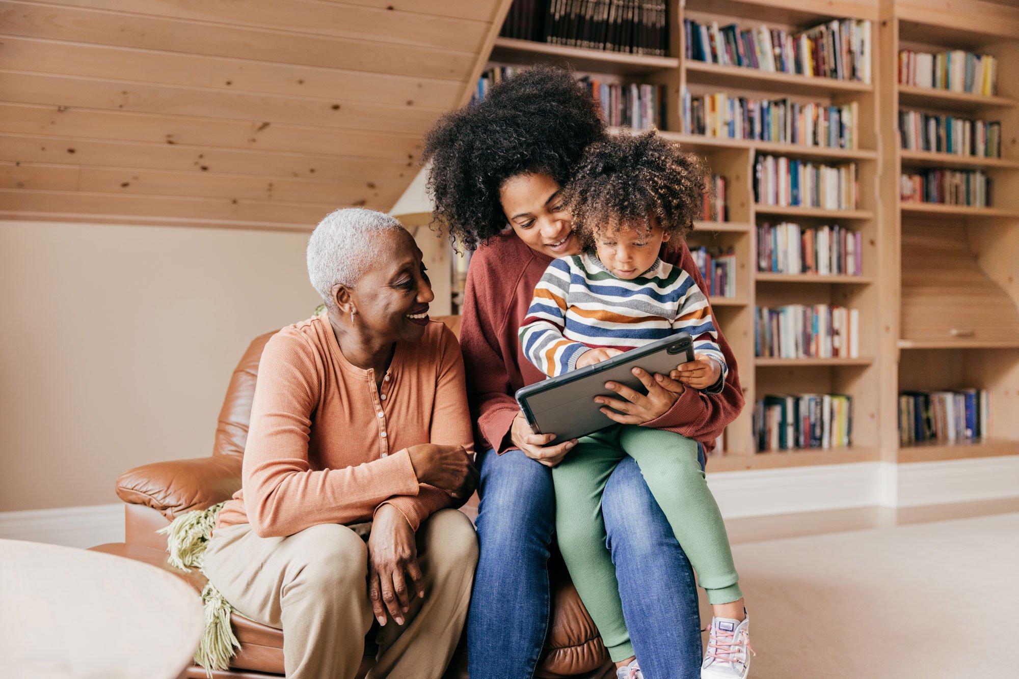 Grandmother, mother, and grandchild sitting together and watching something on a tablet.